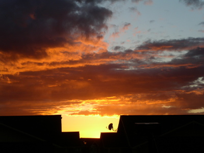 [A satellite dish sticks out from the end of a building and it and the building edges are in a sea of bright yellow. The clouds above that level are bright orange. There is still a patch of blue above the clouds.]